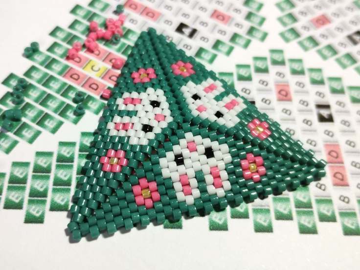 A Peyote triangle with three rabbit heads and pink flowers lying on the bead weaving peyote pattern