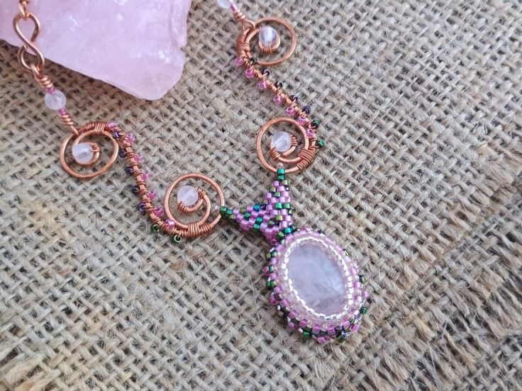 A rose quartz cabochon pendant necklace with wire work and a beaded bezel against a hessian background
